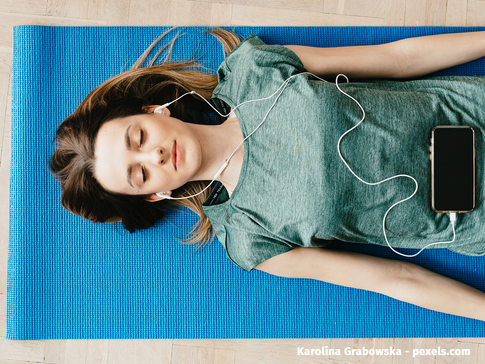 Blick von oben auf eine junge Frau, die mit geschlossenen Augen auf einer blauen Yogamatte liegt. Auf ihrem Bauch liegt ein Smartphone. Über Kopfhörer hört sie sich etwas an.