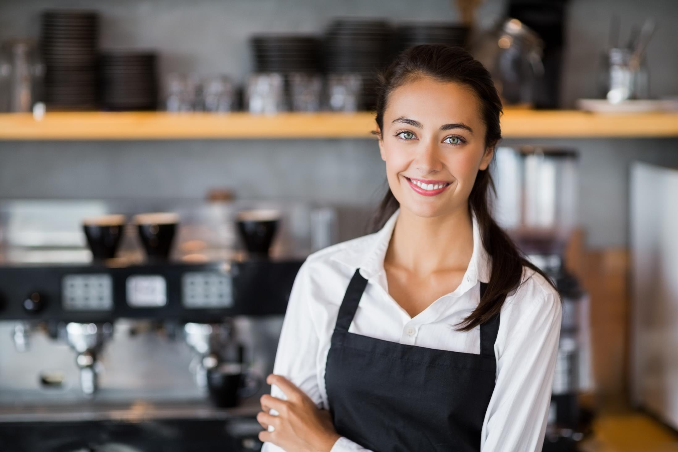 Freundlich lächelnde Mitarbeiterin mit Schürze, im Hintergrund verschwommen ein Café.