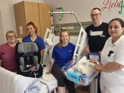 5 Personen stehen um ein Babybett in einem Krankenzimmer. Alle Personen lachen in die Kamera. Ein Kindersitz steht auf dem Bett.