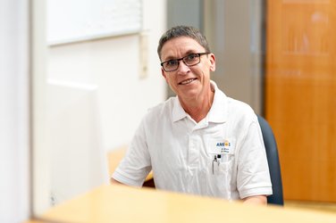 Eine Pflegefachkraft sitzt am Schreibtisch hinter einem Monitor und schaut lächelnd in die Kamera.