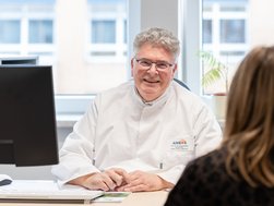 Arzt in Kittel sitzt am Schreibtisch vor einer Patientin und lächelt in die Kamera