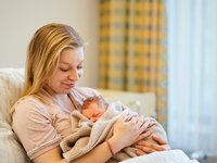 Mutter Leonie Lüder liegt im Bett und hält ihr neugeborenes Baby Lilli im Arm. Sie lächelt es liebevoll an. (© AMEOS).